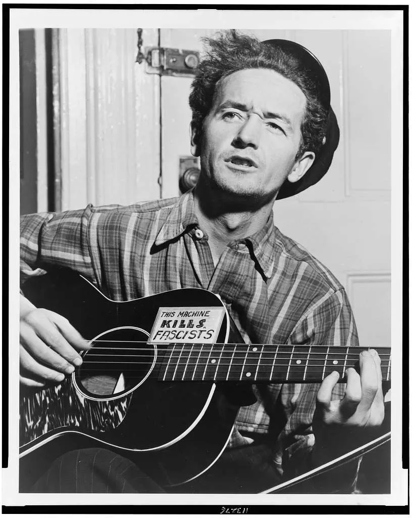 Immagine di Woody Guthrie e la sua chitarra acustica con la scritta "This Machine kills fascists".
